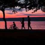 Silhouette of a family walking and biking by a lake at sunset, framed by trees.